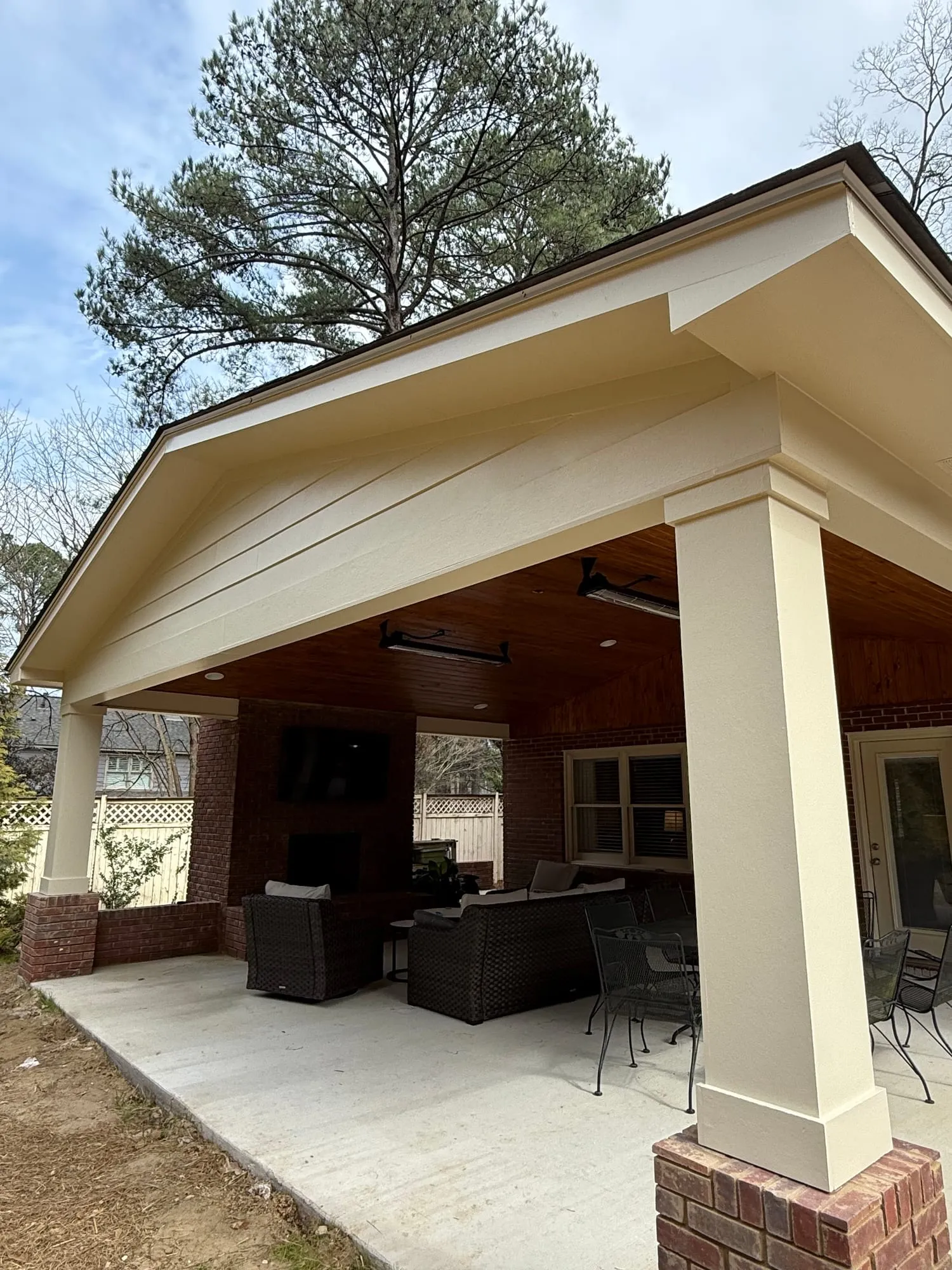 A covered backyard patio with a cream-painted gable and column, a stained tongue-and-groove wood ceiling, a brick fireplace, and wicker seating on a concrete slab.