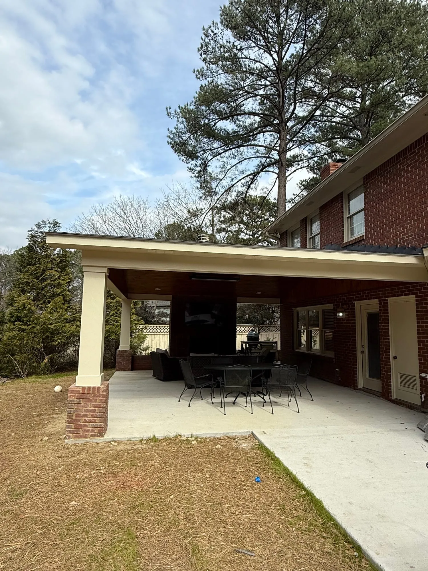 A covered patio attached to a two-story brick home, with a cream-painted beam and column, a dining set underneath, and a lawn out front.