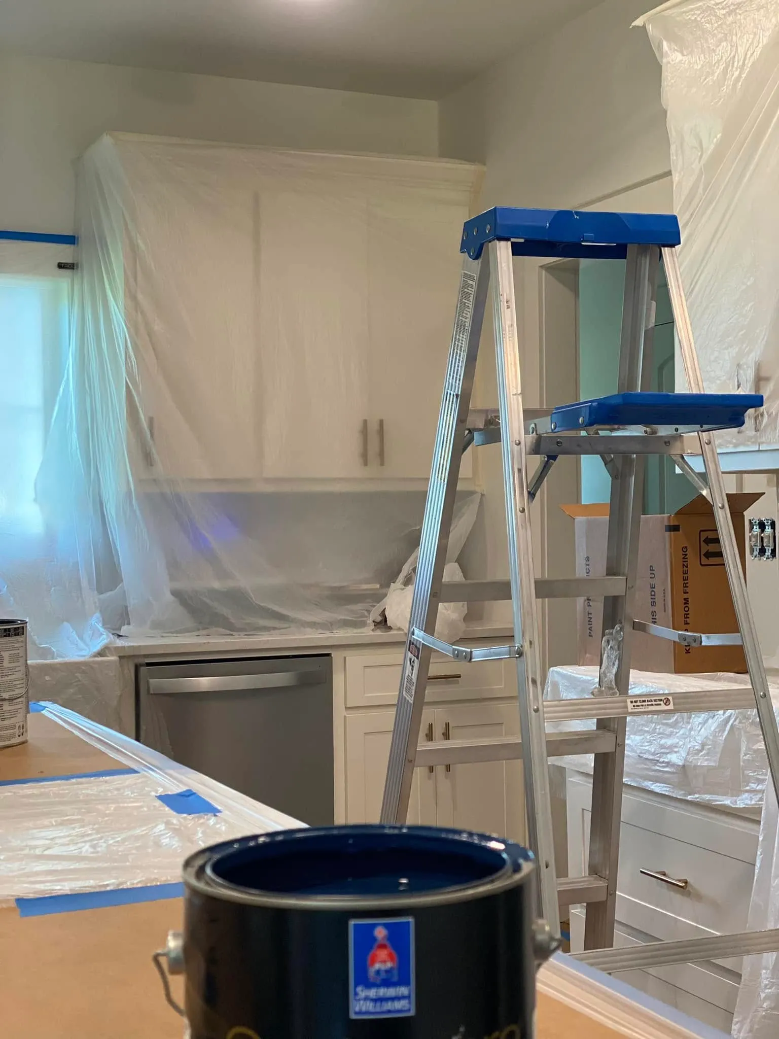 A kitchen fully masked in plastic sheeting and blue tape with a step ladder and a Sherwin-Williams paint can, ready for painting.