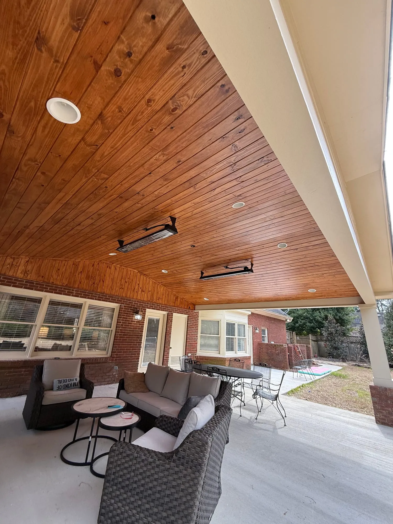 A stained wood patio ceiling meeting a brick wall with paned windows, recessed lights and heaters above a wicker seating group.