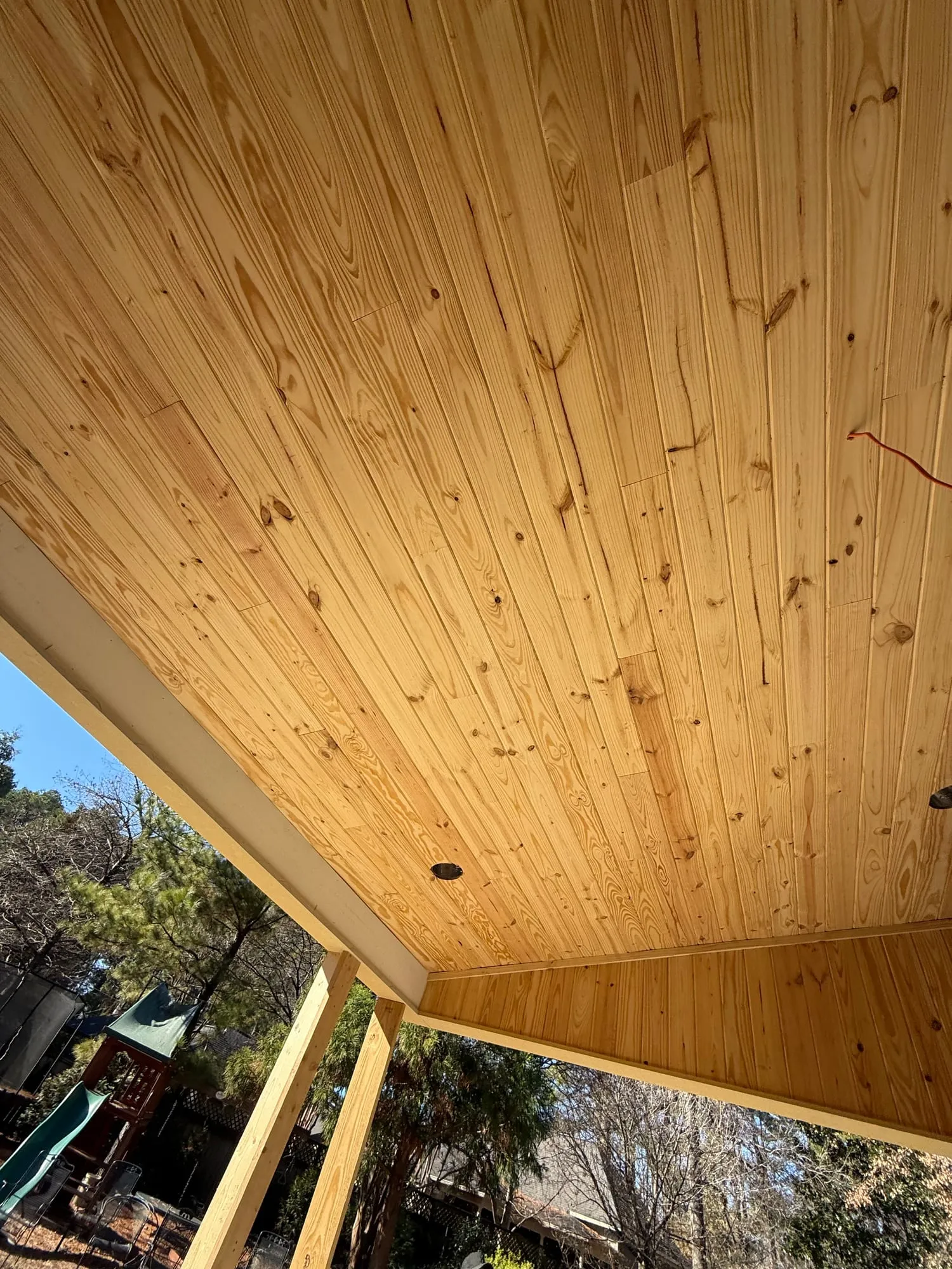 A newly installed patio ceiling of bare, unfinished pine tongue-and-groove boards, with trees and blue sky past the roof edge.
