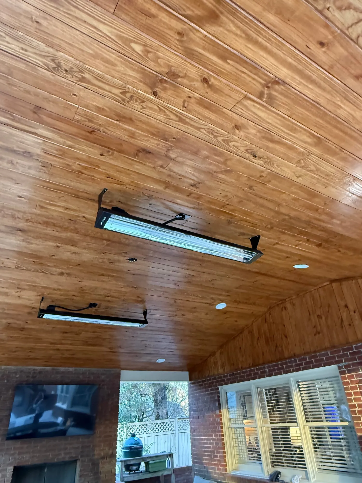 A high vaulted pine patio ceiling in a rich golden stain, with patio heaters and recessed downlights, over a brick fireplace and a kamado grill.