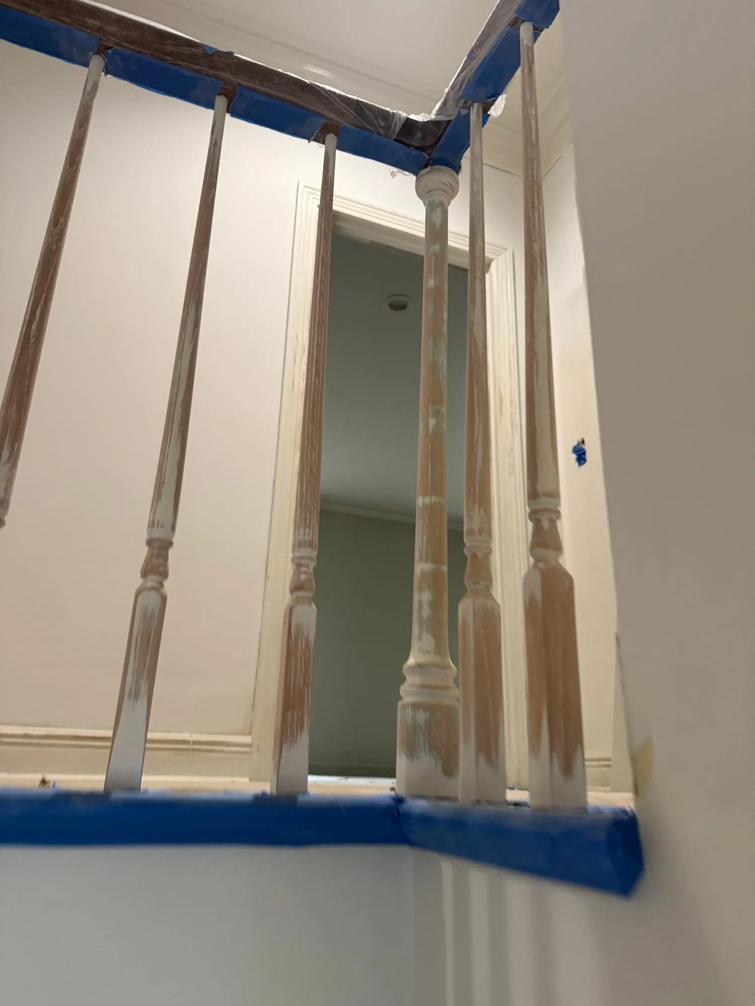 A staircase railing with turned wood balusters primed in white, blue tape masking the handrail and floor, looking through a doorway into a sage room.